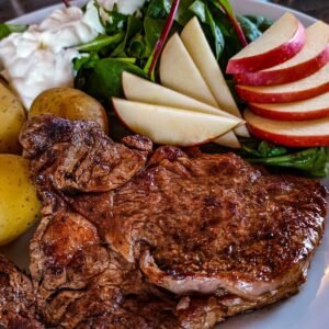 Delicious grilled steak with potatoes, fresh salad, and apple slices on a white plate.